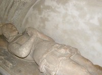 The effigy of a man in a tunic on a table tomb, his hands together in prayer.